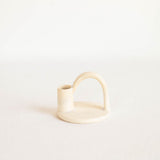 A minimalist ceramic candle holder with a flat circular base, a cylindrical cup, and a curved handle, all in a warm cream or off-white glaze, set against a plain white background.