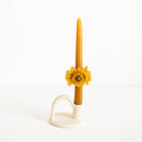 A single taper candle in a warm golden-yellow color, decorated with a realistic yellow flower at the center, resting in a cream-colored ceramic holder against a plain white background.