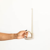 A hand holding a minimalist, modern candle holder with a single tall taper candle against a solid white background.