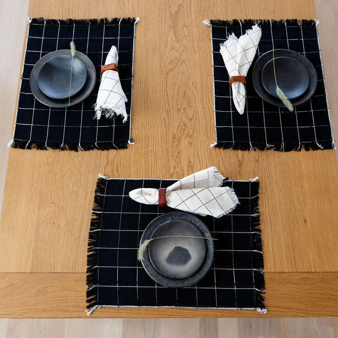 Three black cotton woven placemats with white checkered patterns on a wooden dinner table.