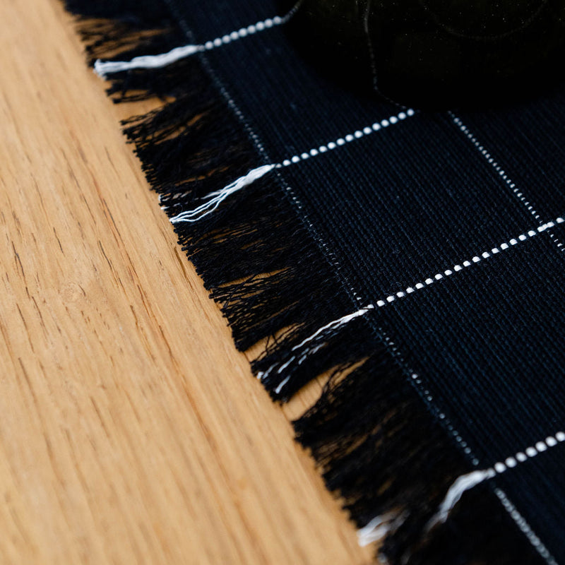 Close-up of a black and white checkered cotton placemat with frayed edges on a wooden dinner table.