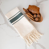 Beige Mexican cotton hand towel with hunter green stripes and hand-applied fringe against a marble background and Mexican pan dulce bread on the side