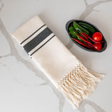 Beige Mexican cotton hand towel with black stripes and hand-applied fringe against a marble background and jalapeno peppers and tomatoes on the side
