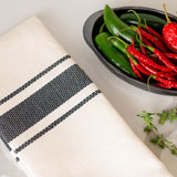 Close up of a beige Mexican cotton hand towel with black stripes with a tray of jalapeno peppers and tomatoes on the side