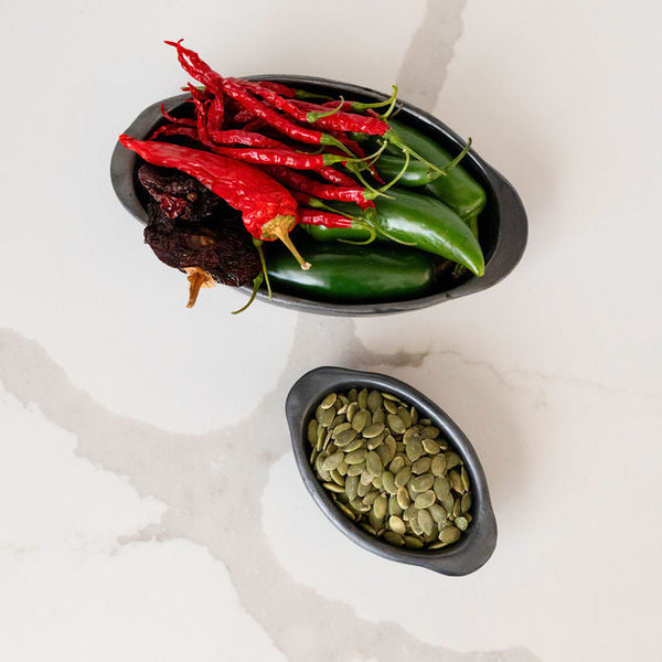 Two black ceramic serving bowls containing red and green peppers on a light background