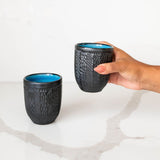 Two black textured Mexican ceramic cups with blue interior on a white marble surface.