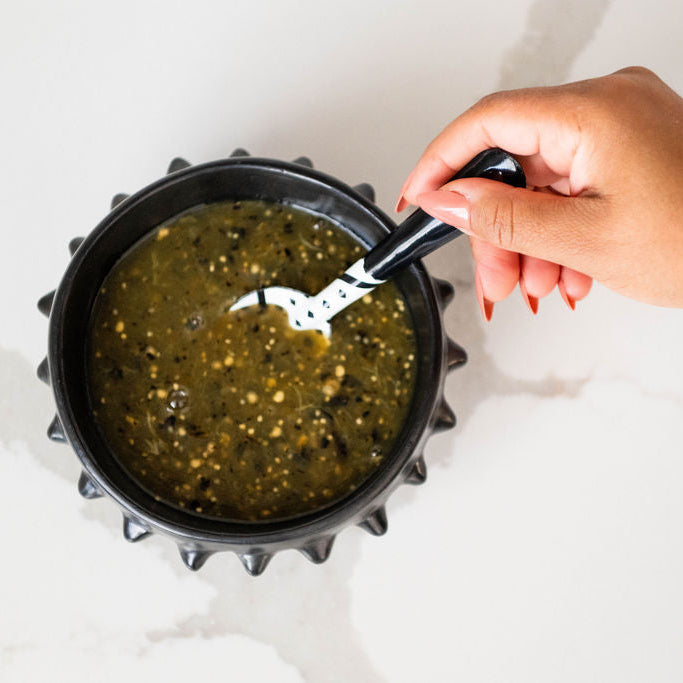 Hand stirring a salsa verde inside a black ceramic serving bowl on a white background