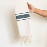 Hand holding a natural Mexican cotton hand towel with hunter green stripes and hand-applied fringe against a plain background