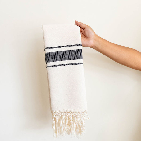 A hand holding a hand-woven cotton beige hand towel with black stripes and fringe