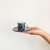 Hand holding a black and white patterned expresso mug and saucer against a plain background