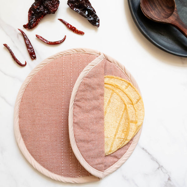 Round cotton woven tortilla warmer in cinnamon color on a white surface with dried chilies and a wooden spoon.