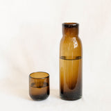 Chocolate brown tall glass carafe with water inside and a lid that acts as a cup with white background