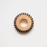 A close-up of a circular wooden napkin ring with a distinctive striped pattern. The ring is made of light wood (pinewood) with black/dark stripes evenly spaced around its circumference, creating a cogwheel-like appearance. The wooden ring has a smooth, polished finish with a hollow center.