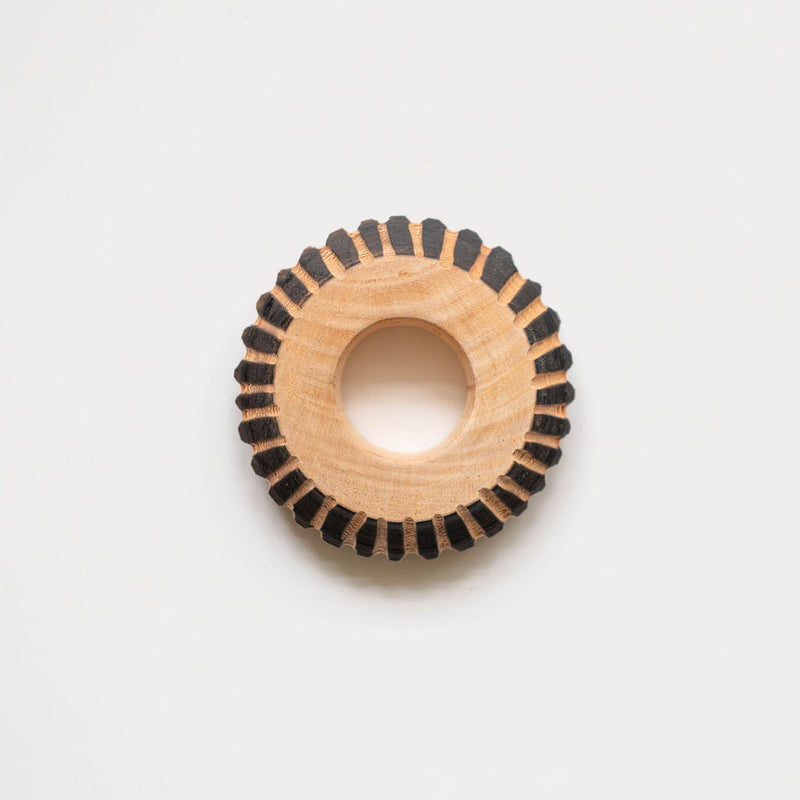 A close-up of a circular wooden napkin ring with a distinctive striped pattern. The ring is made of light wood (pinewood) with black/dark stripes evenly spaced around its circumference, creating a cogwheel-like appearance. The wooden ring has a smooth, polished finish with a hollow center.