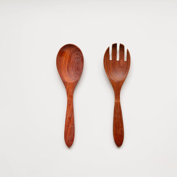 A minimalist product photo showing a wooden serving set against a white background. The set consists of a smooth, reddish-brown wooden spoon on the left and a matching wooden serving fork with three tines on the right. Both utensils display the natural wood grain and warm color of Mexican Rosewood.