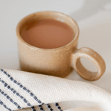 Large Handle Ceramic Stoneware Mug - Brown / Nuez / beige color with coffee or tea inside and a napkin on a counter