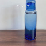 Video showing water pouring from a handblown blue glass bottle onto a cup that serves as a lid on a brown table