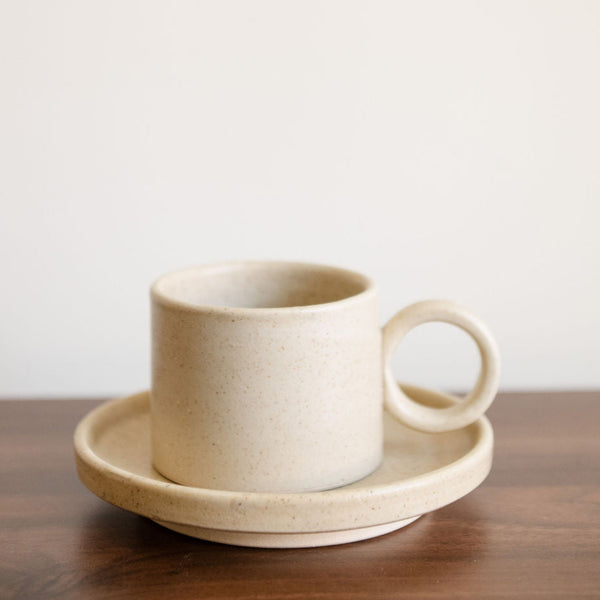 Vanilla Creme Mexican Ceramic Cup and Saucer Set featuring a wide ear handle and cream-colored finish with subtle speckles and matching saucer plate sitting on a brown dinner table. Perfect for serving coffee or tea, this set combines simplicity and elegance.