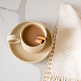 Vanilla Beige Mexican Ceramic Mug and saucer with wide ear handle filled with coffee and cinnamon sticks on a marbled kitchen counter.